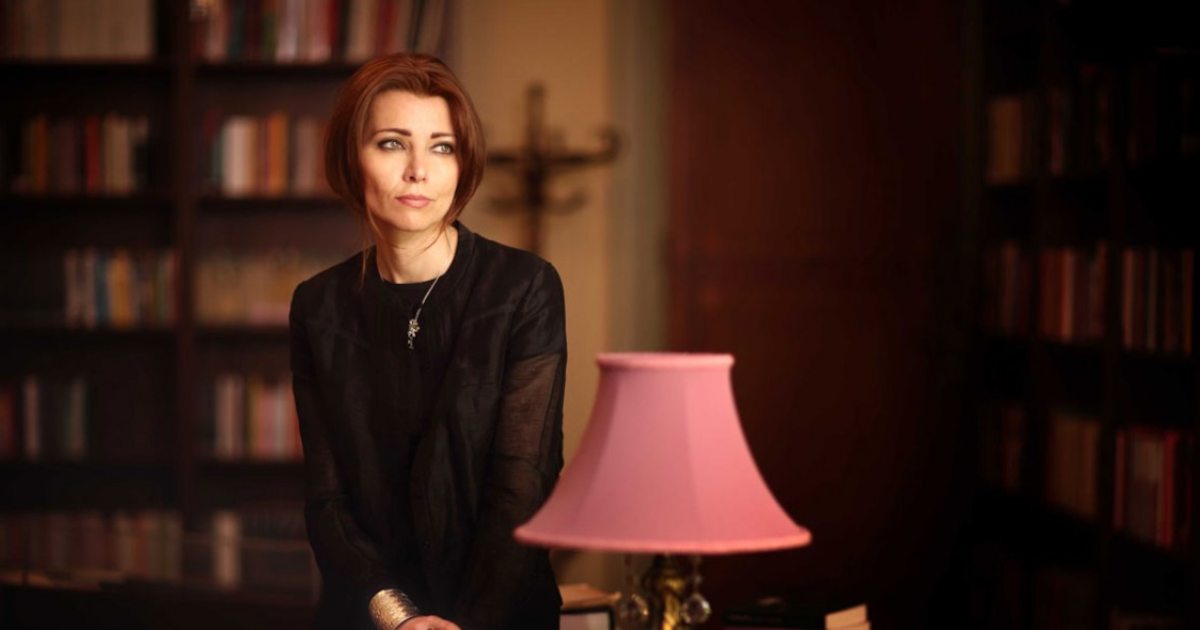 Elif Şafak sitting on a desk with bookshelves in the background and a lamp with a pink shade in the foreground